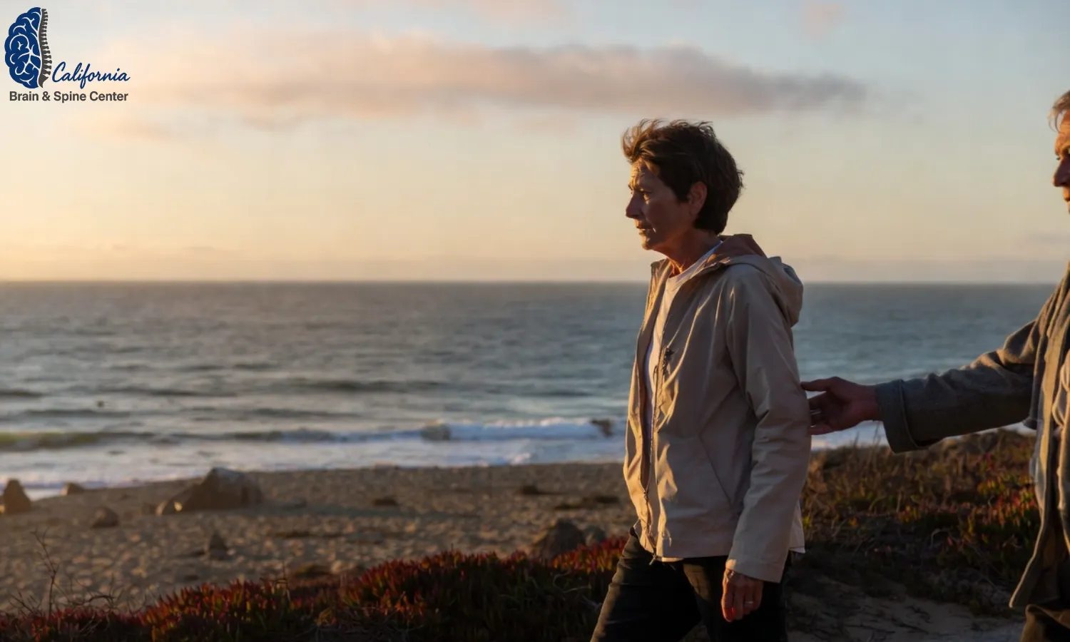A middle aged person walking slowly along a Malibu shoreline path, looking cautious but hopeful, with subtle support from a family member, soft sunset lighting, calm mood.”